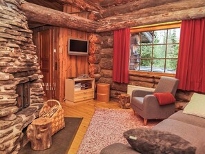 Interior de una cabaña de troncos con una pared de piedra, un televisor montado en la pared, una chimenea, un armario de madera, una ventana con cortinas rojas y un rincón de estar acogedor con un sofá y un sillón.