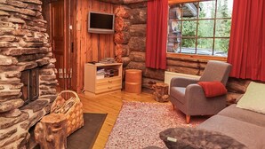 Een houten blokhut van binnen, met een stenen muur, een open haard, een televisie en een raam met rode gordijnen.