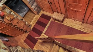 Een houten blokhut van binnen met een open haard, een houten bank en een vloerkleed met rode en bruine strepen.
