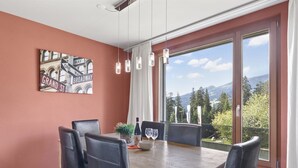Un coin repas avec une table en bois, des chaises en cuir noir, une bouteille de vin et une vue sur les montagnes, visible à travers de grandes fenêtres.