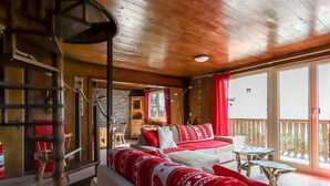 L'interno accogliente di un rifugio con soffitto in legno, un divano con cuscini rossi e bianchi, un tavolo da pranzo e un balcone con ringhiera in legno.