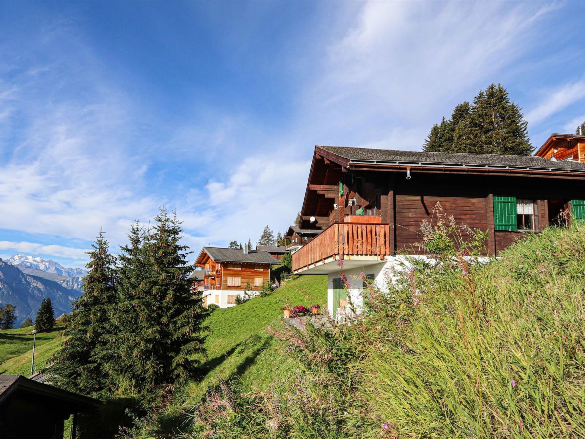 Eine Holzchalet mit Balkon, umgeben von üppiger Vegetation und Bergen im Hintergrund.