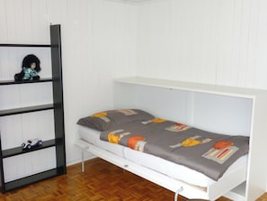 Une petite chambre blanche, avec un lit simple, un plancher en bois, une étagère intégrée et un lit recouvert d’une couvre-lit à motifs gris et orange.