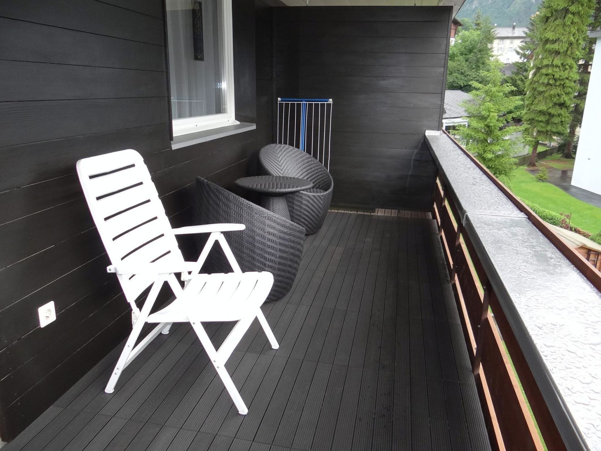 A balcony with a white chair and a round table, a window, and a railing overlooking a green landscape.