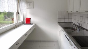 Una cocina con armarios blancos, una cafetera roja, un ventanal con cortinas de encaje y vistas al exterior con vegetación.