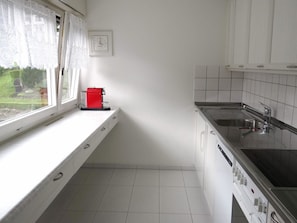 Una cucina con mobili bianchi, una macchina da caffè rossa, una finestra con tende in pizzo e vista su del verde all'esterno.