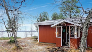 Pieni puinen mökki, jonka ovi on auki, sijaitsee vesistön lähellä ja sen ympärillä on puita.