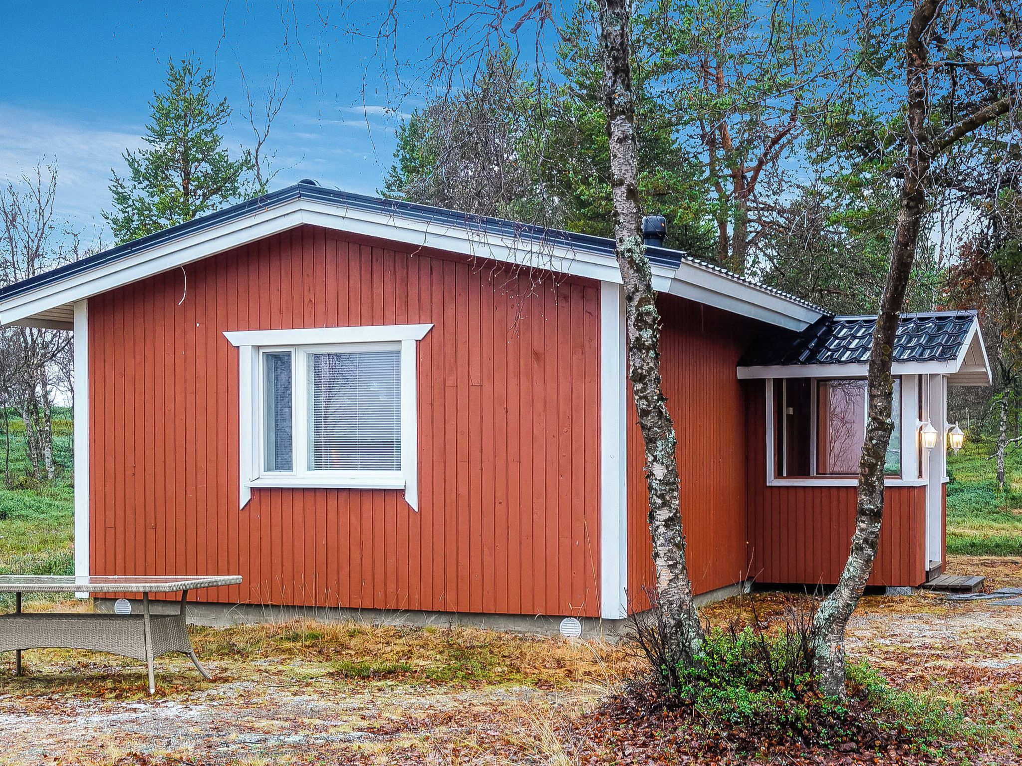 A red wooden cabin with a single window and a small attached structure, surrounded by trees and a grassy area.