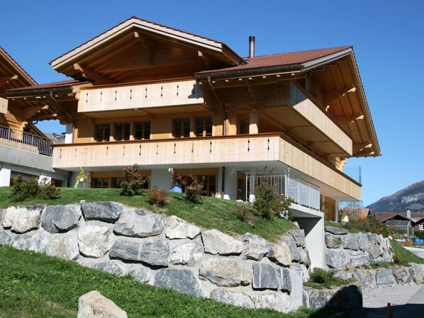 Un edificio de madera de varios pisos con balcones, situado en una colina con un muro de contención de piedra.