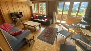 A spacious living room with a wooden ceiling, a flat-screen TV, a sofa, a coffee table, and a view of a green landscape through large windows.