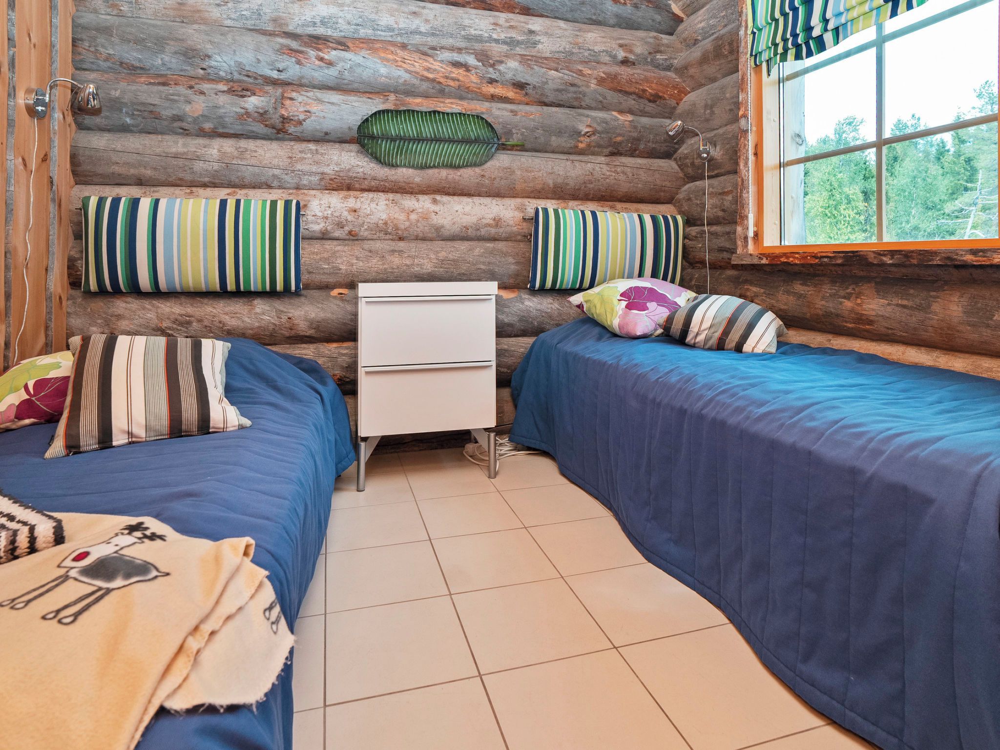 A log cabin room with two beds, a small white dresser, and a window with a view of trees.