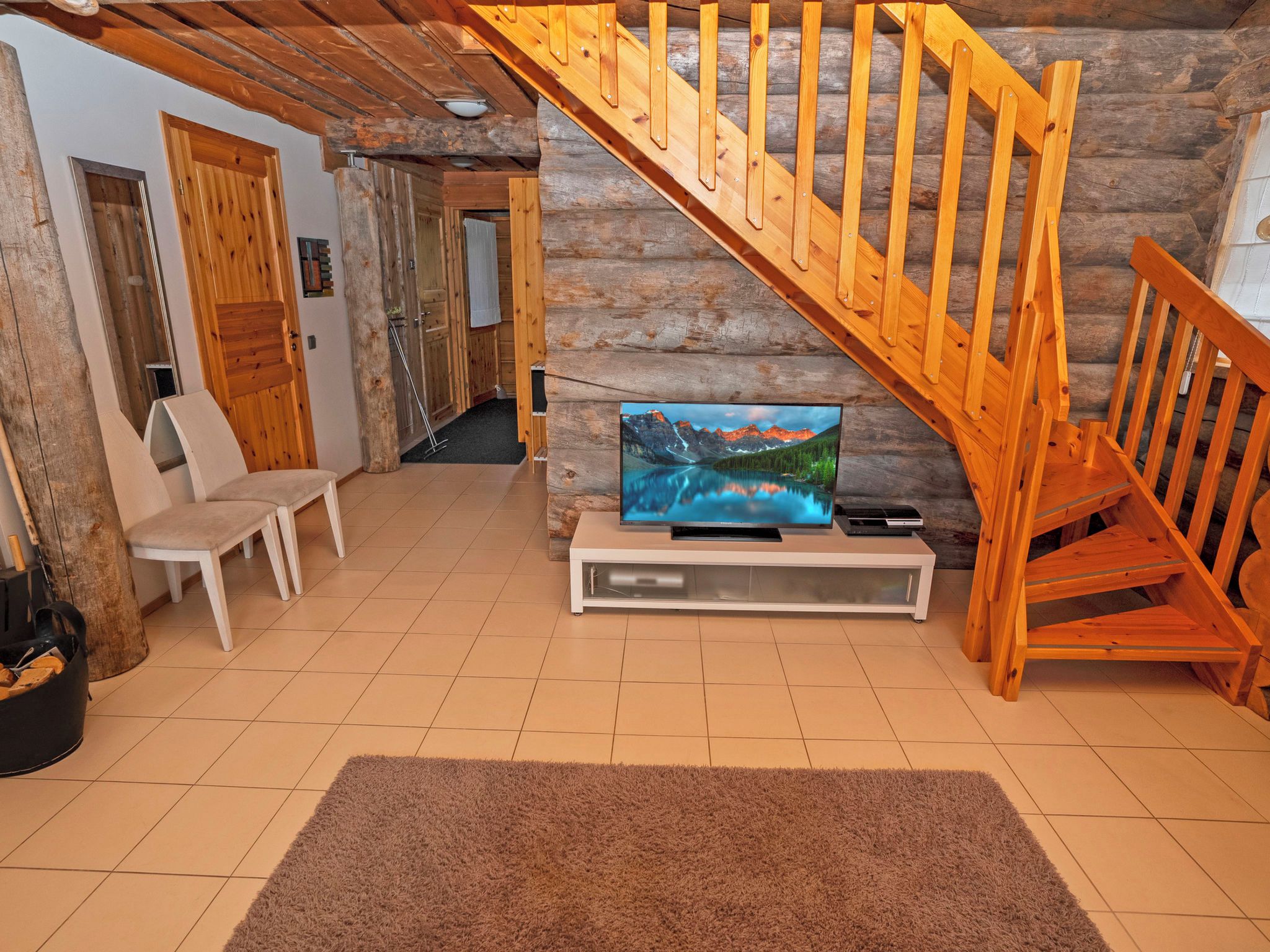 A log cabin interior with a wooden staircase, a television on a white stand, a bench, and a fireplace.