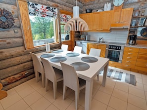 L’intérieur d’une cabane en rondins comprend une table à manger, des chaises, un luminaire suspendu, des armoires en bois, un coin cuisine et une fenêtre donnant sur les arbres.