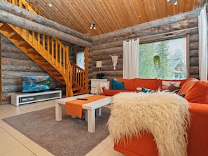 L’intérieur d’une cabane en rondins, avec un escalier en bois, un téléviseur, un canapé et une grande fenêtre équipée de stores.