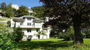 Une maison à deux étages avec un balcon, entourée de verdure et d’un grand arbre.