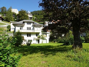 Une maison à deux étages avec un balcon, entourée de verdure et d’un grand arbre.