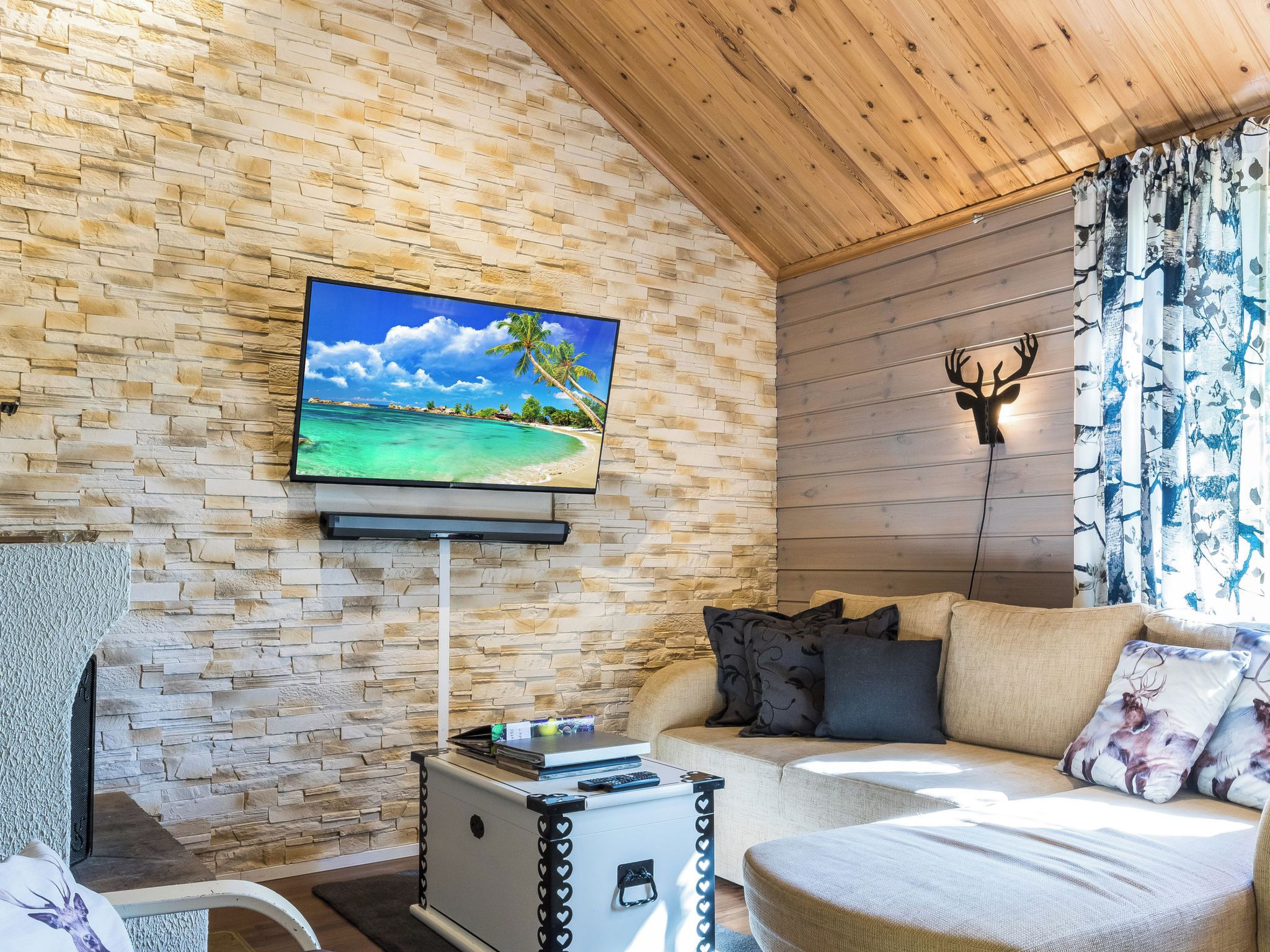 Un salon chaleureux avec un mur en pierre, un téléviseur à écran plat, un plafond en bois et un canapé agrémenté de coussins décoratifs.