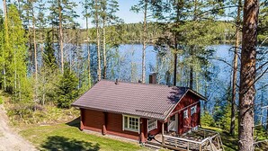 A cabin with a red exterior, a chimney, and a porch with a swing set, surrounded by trees and a lake.