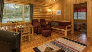 A cozy log cabin interior with a wooden dining table, red upholstered chairs, a sofa, and a rug.