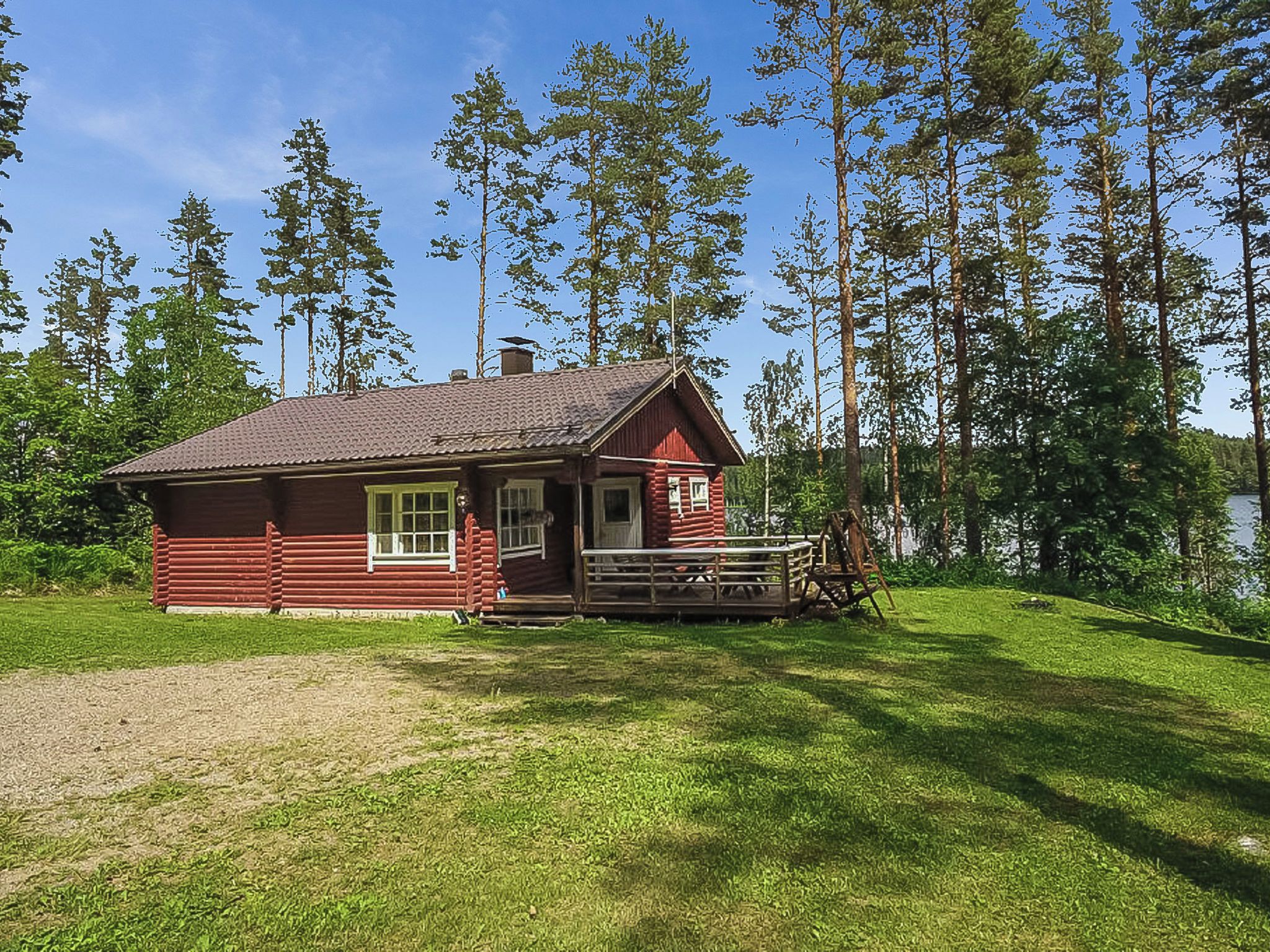 Punainen hirsihökkelö, jossa on kuisti ja puinen keinutuoli, ja sen ympärillä on puita ja ruohikkoista aluetta.