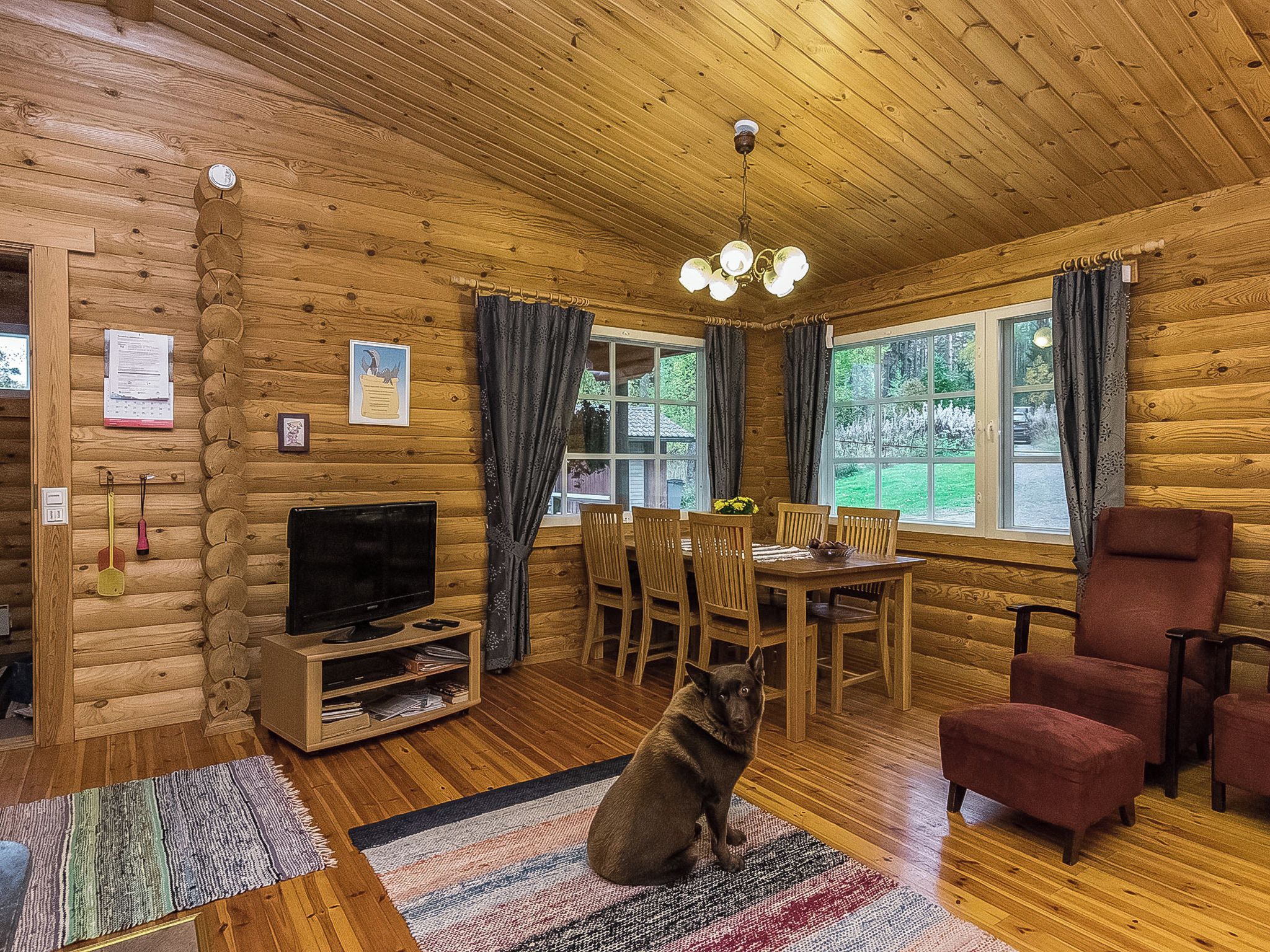 A log cabin interior with a wooden floor, a television, a dining table with chairs, and a dog sitting on a rug.
