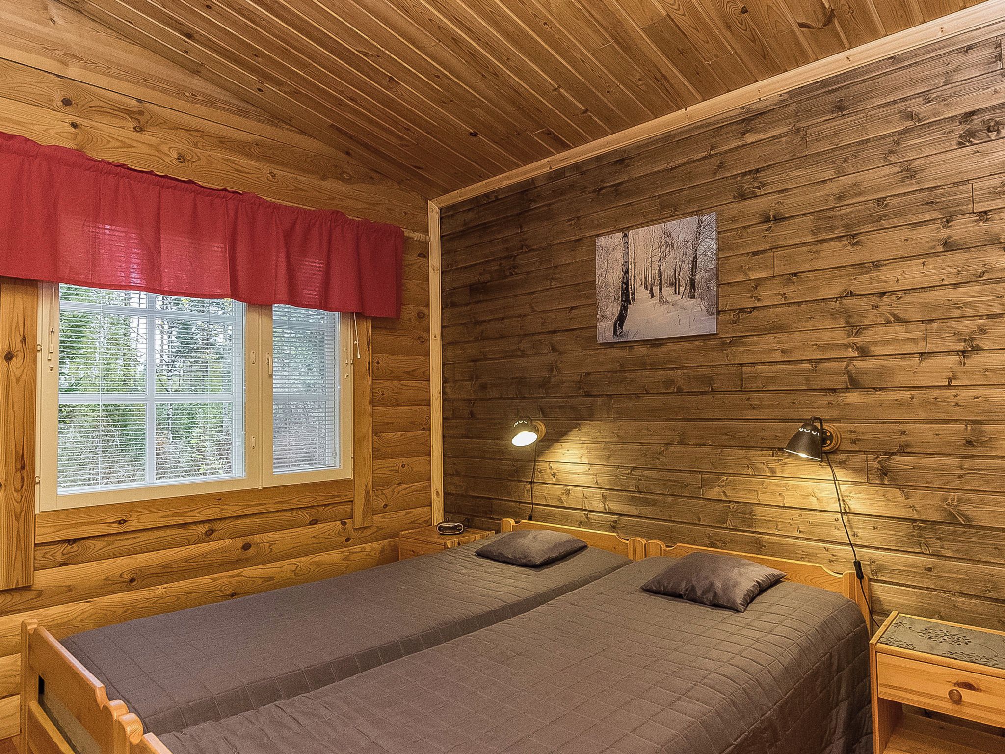 A wooden cabin room with a bed, two bedside lamps, a window with blinds, and a painting on the wall.