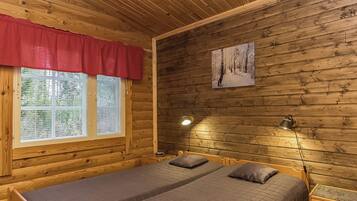 A wooden cabin room with a bed, two bedside lamps, a window with blinds, and a painting on the wall.