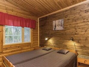 A wooden cabin room with a bed, two bedside lamps, a window with blinds, and a painting on the wall.