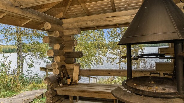 A rustic outdoor seating area with a wood-burning stove, benches, and a view of a lake.