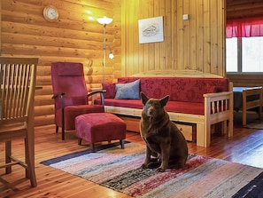 A cozy cabin interior with a wooden floor, a red sofa, a red chair, a wooden bench, and a dog sitting on a rug.