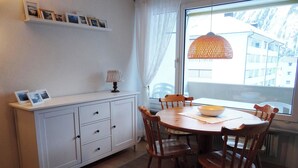 A dining area with a round table, wooden chairs, a white cabinet, and a window with a view of buildings. - Neuschwändi 63 Appartement/Fewo 4-Bettwohnung by Interhome (Engelberg)