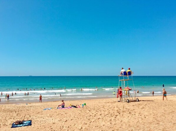 A beach with lifeguards on a tower, people swimming and walking on the sand, and a clear blue sky.