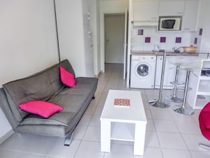 A modern living room with a grey sofa, a white coffee table, and a red chair. A washing machine and a microwave are visible in the background.