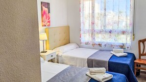 Camera d'albergo con due letti, un comodino, una lampada, una finestra con tende floreali e una lampada a sospensione blu.