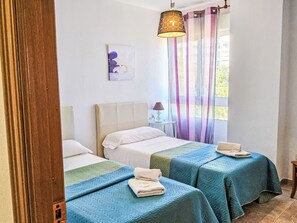 Een kamer met twee bedden, een raam met gordijnen, een hanglamp en een klein tafeltje met lamp.