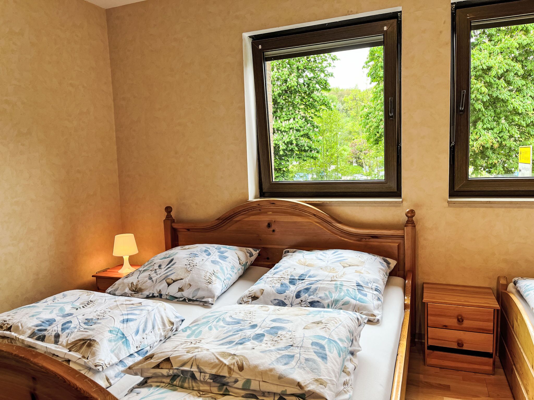Une chambre à coucher avec un lit en bois, deux fenêtres donnant sur un paysage verdoyant, une table de chevet avec une lampe et une petite table de nuit en bois.