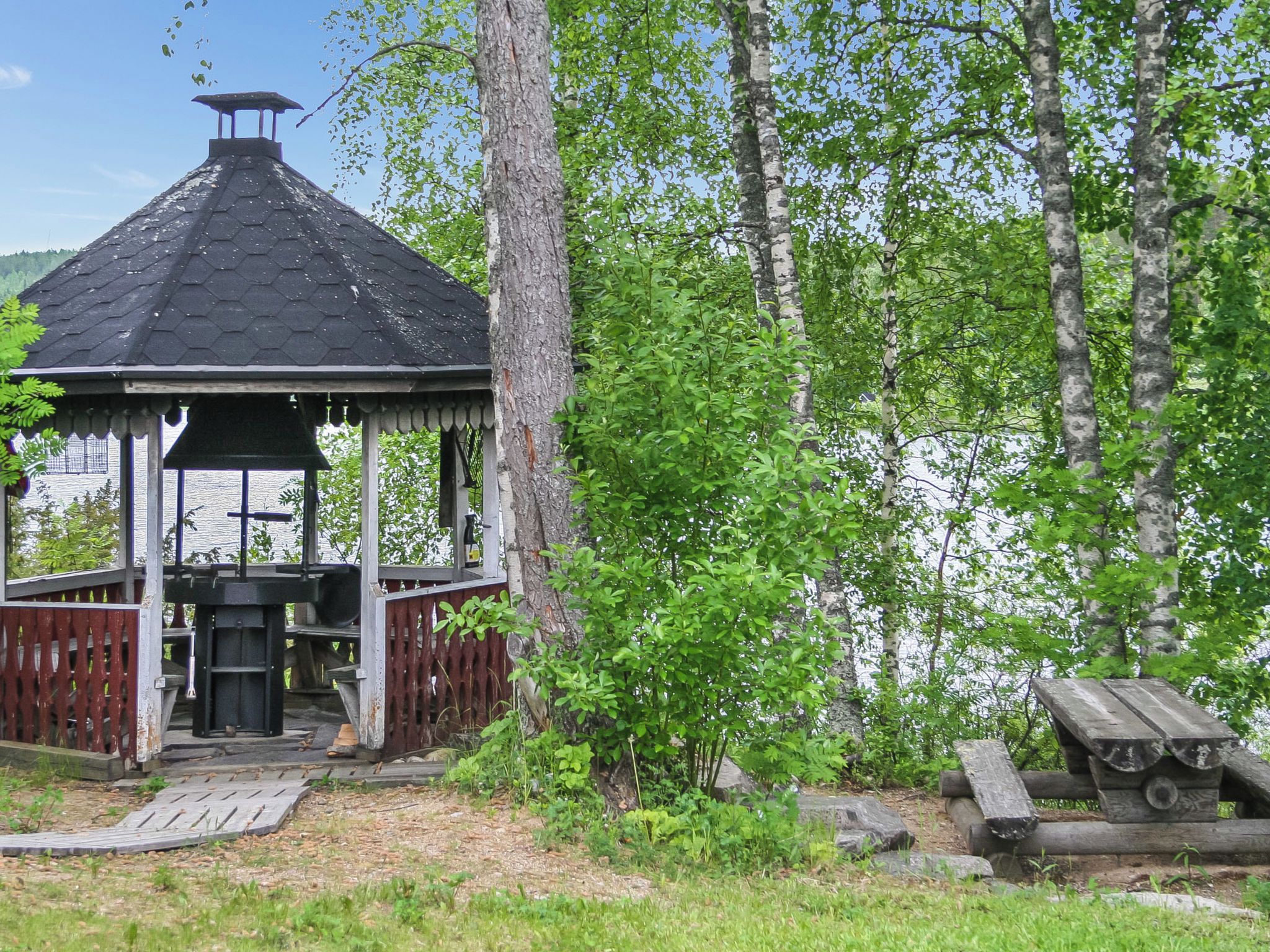 Kuusikulmaisella katolla varustettu huvimaja, jonka ympärillä on puinen terassi, puita ja piknikpöytä.