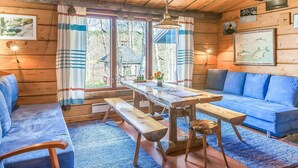 Een gezellig chaletinterieur met een houten plafond, een eettafel met banken, een blauwe bank en een groot raam met gordijnen.