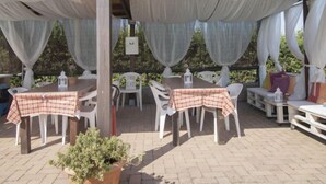 Eine Terrassengruppe mit Tischen und Stühlen, überdacht mit einem Sonnensegel, umgeben von viel Grün.
