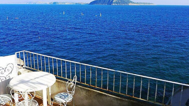 Un balcon avec une table et des chaises donnant sur une mer d’un bleu profond, avec une île visible au loin.