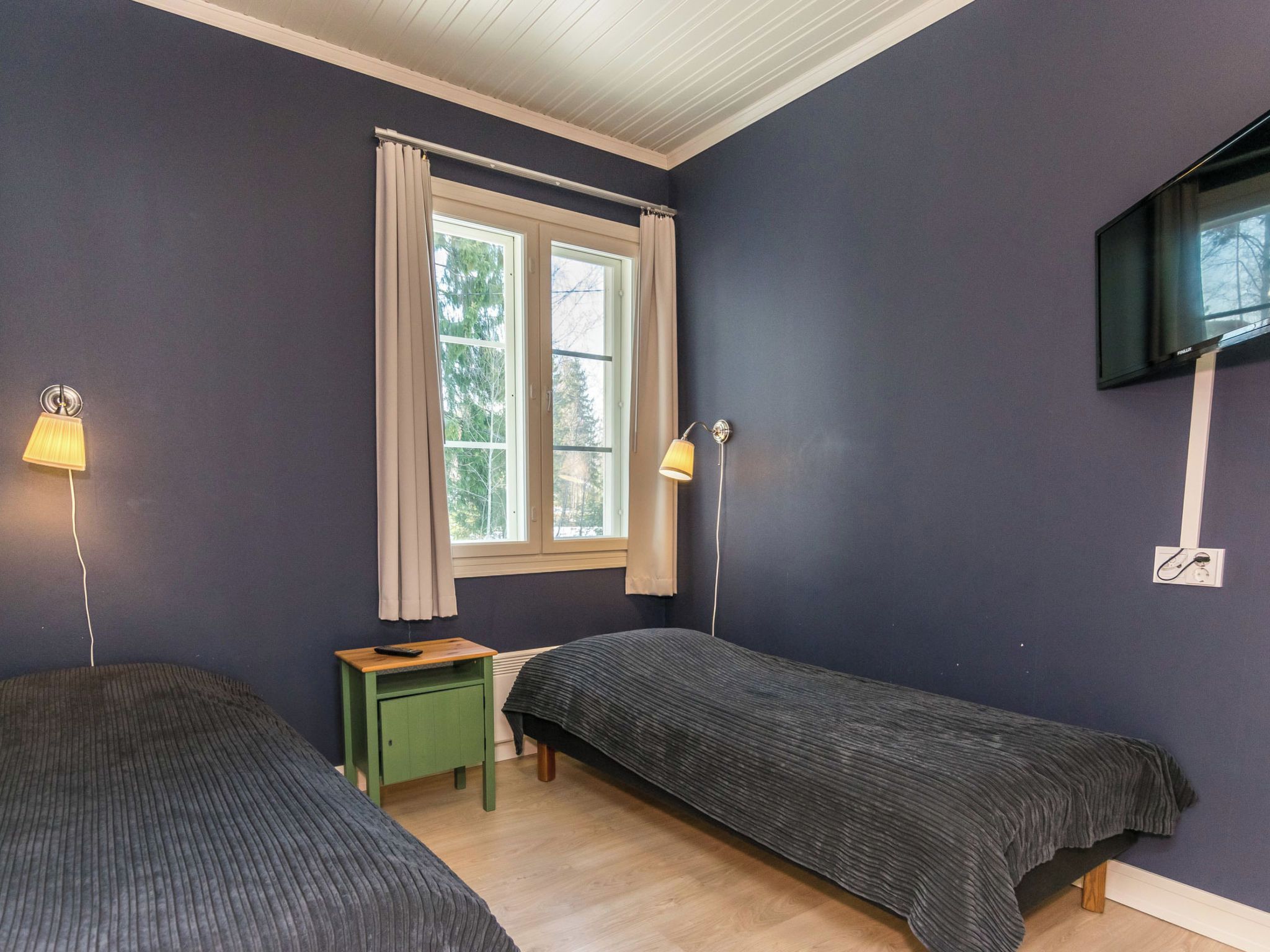Ein Zimmer mit zwei Betten, einem fensterbelaubten Fenster, einem kleinen Tisch, einer Lampe und einem an der Wand befestigten Fernseher.