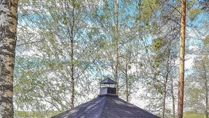 A wooden gazebo with a sloped roof and a chimney, situated by a lake surrounded by trees. - Männikkö by Interhome (Sulkava)