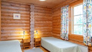 A log cabin room with two beds, a wooden wall, a window with curtains, and a hanging light fixture. - Männikkö by Interhome (Sulkava)