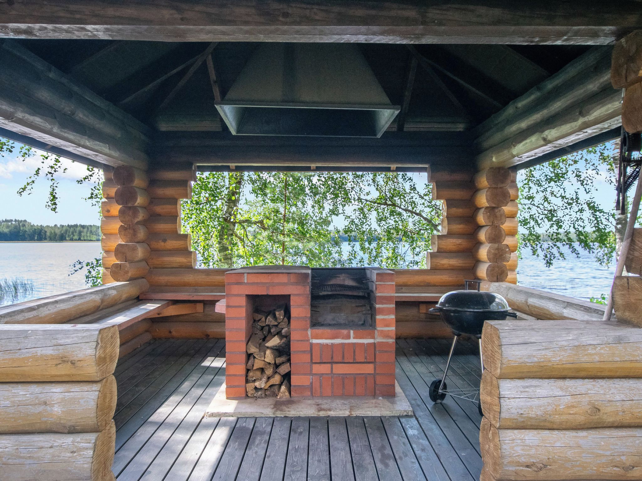 A log cabin with a brick fireplace and a grill, overlooking a lake.