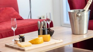 Una mesa de madera con una tabla de cortar, queso, un tarro de miel, una botella de aceite de oliva, una copa de vino, una botella de champán y un cubo de metal con un abrelatas.