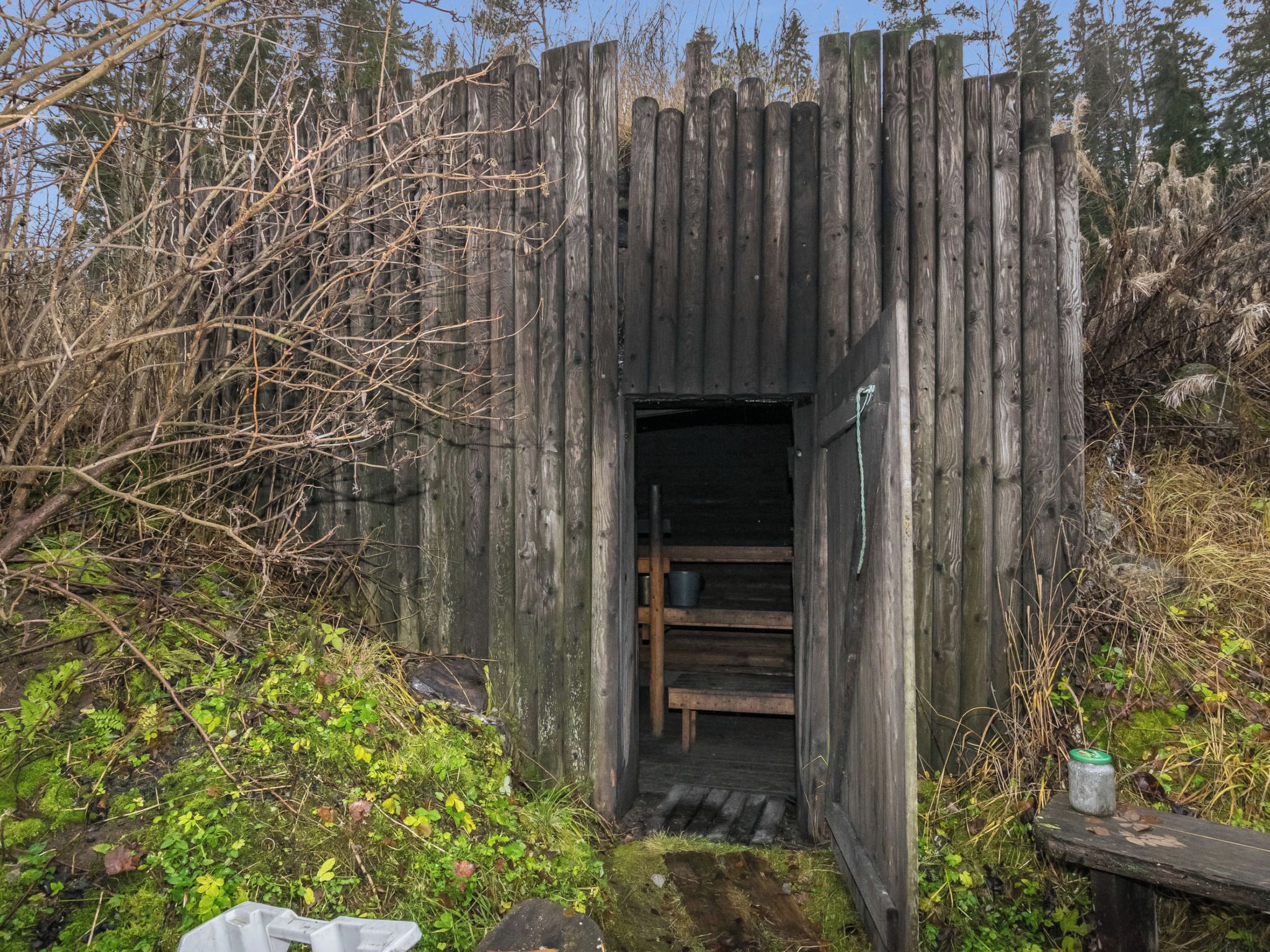 Luonnonmukaisesta puusta rakennettu, maalaishenkinen rakennelma, jonka sisällä on penkki ja jonka ympärillä kasvaa luonnonkasvillisuutta.