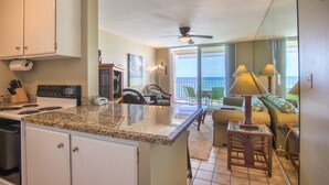 A kitchen with a granite countertop, white cabinets, and a built-in oven. There is a dining area with a table and chairs, and a balcony with a view of the ocean.
