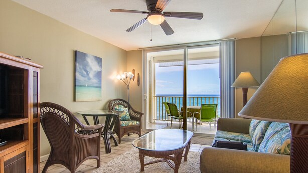 A spacious living area with a ceiling fan, a television, wicker furniture, a glass table, and a view of the ocean through large windows.