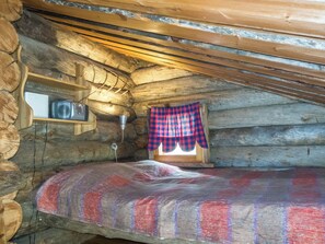 Une chambre confortable dans un chalet en rondins, avec un lit, une petite étagère sur laquelle repose un appareil photo et une fenêtre ornée d’un rideau à carreaux.