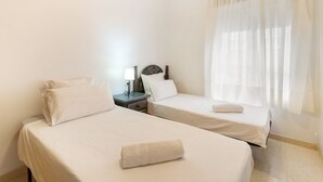 Ein Hotelzimmer mit zwei Einzelbetten, einem Nachttisch mit Lampe und einem Fenster mit durchsichtigen Vorhängen.